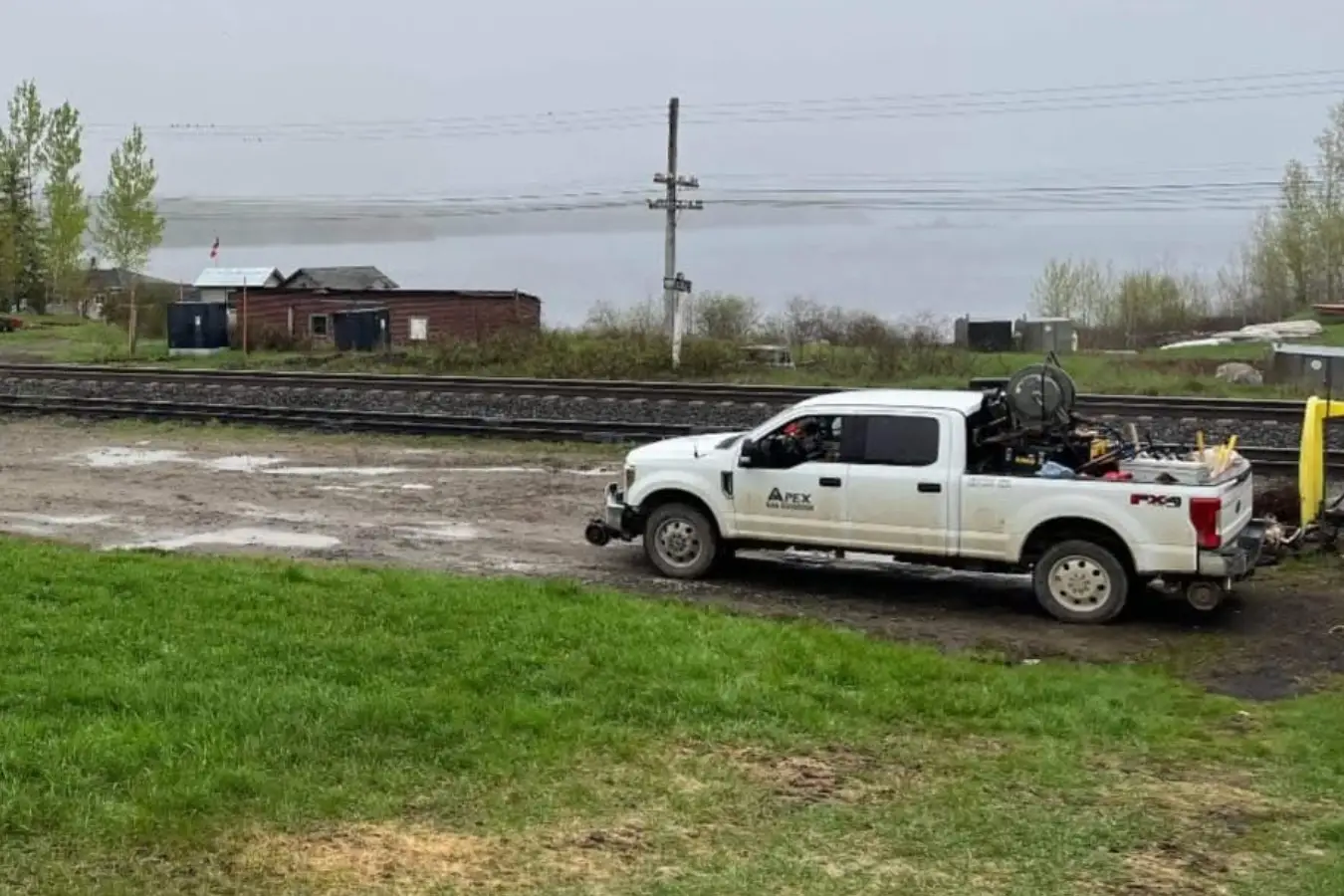 A white truck parked near train tracks with equipment in the bed, beside a grassy area and buildings in a foggy landscape.