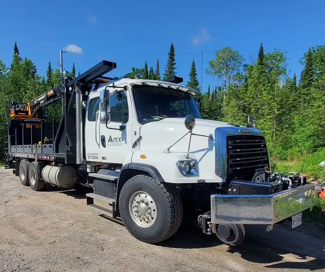A white utility truck with a black flatbed and crane attachment parked on a gravel road, surrounded by lush green trees.