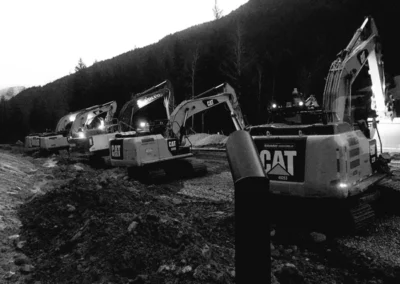 A line of CAT excavators work in a dimly lit construction site amid a mountainous backdrop, showing a busy machinery setup.