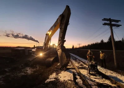 A construction crew works on a railway at dusk, with a large excavator, smoke billowing from a nearby train, and power lines in view.