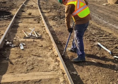 A worker in a safety vest and helmet uses a tool on railroad tracks at a construction site, surrounded by debris and machinery.