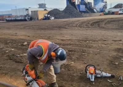 A worker in safety gear cuts metal rail with a power saw, sparks flying, beside tools on a gravel site with machinery in the background.