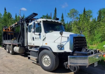 A white utility truck with a black flatbed and crane attachment parked on a gravel road, surrounded by lush green trees.