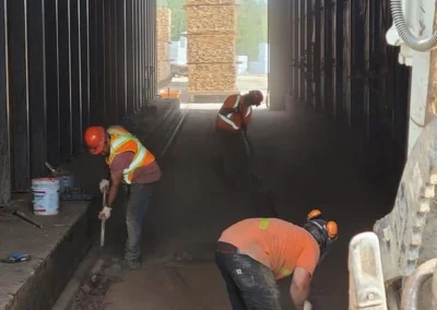 Three construction workers in helmets and vests are cleaning a dusty tunnel with stacks of wood visible in the background.